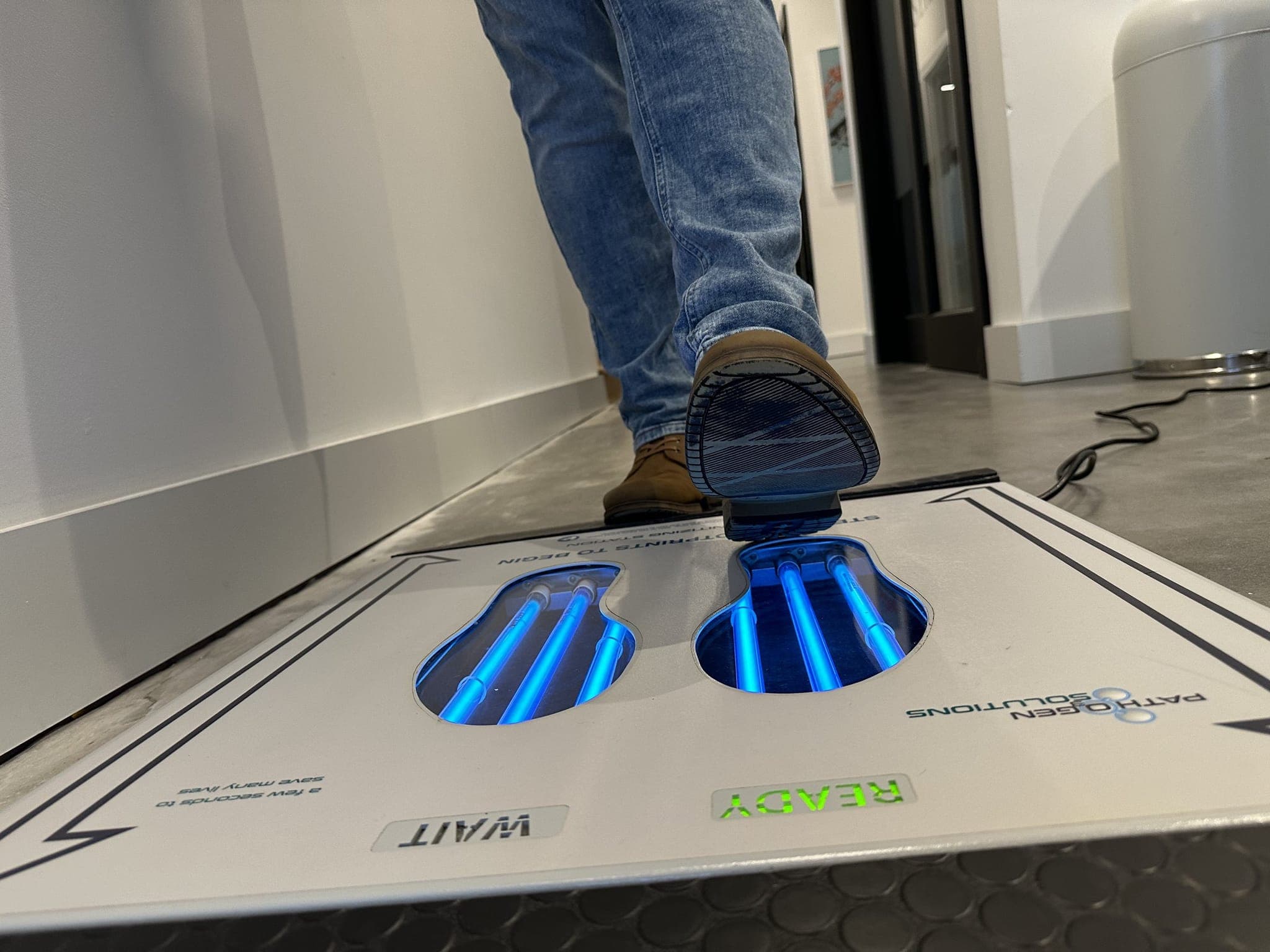 Person in jeans and brown boots stepping across the UVZone shoe sanitizing station, seen from floor level at one end with the READY indicator lit