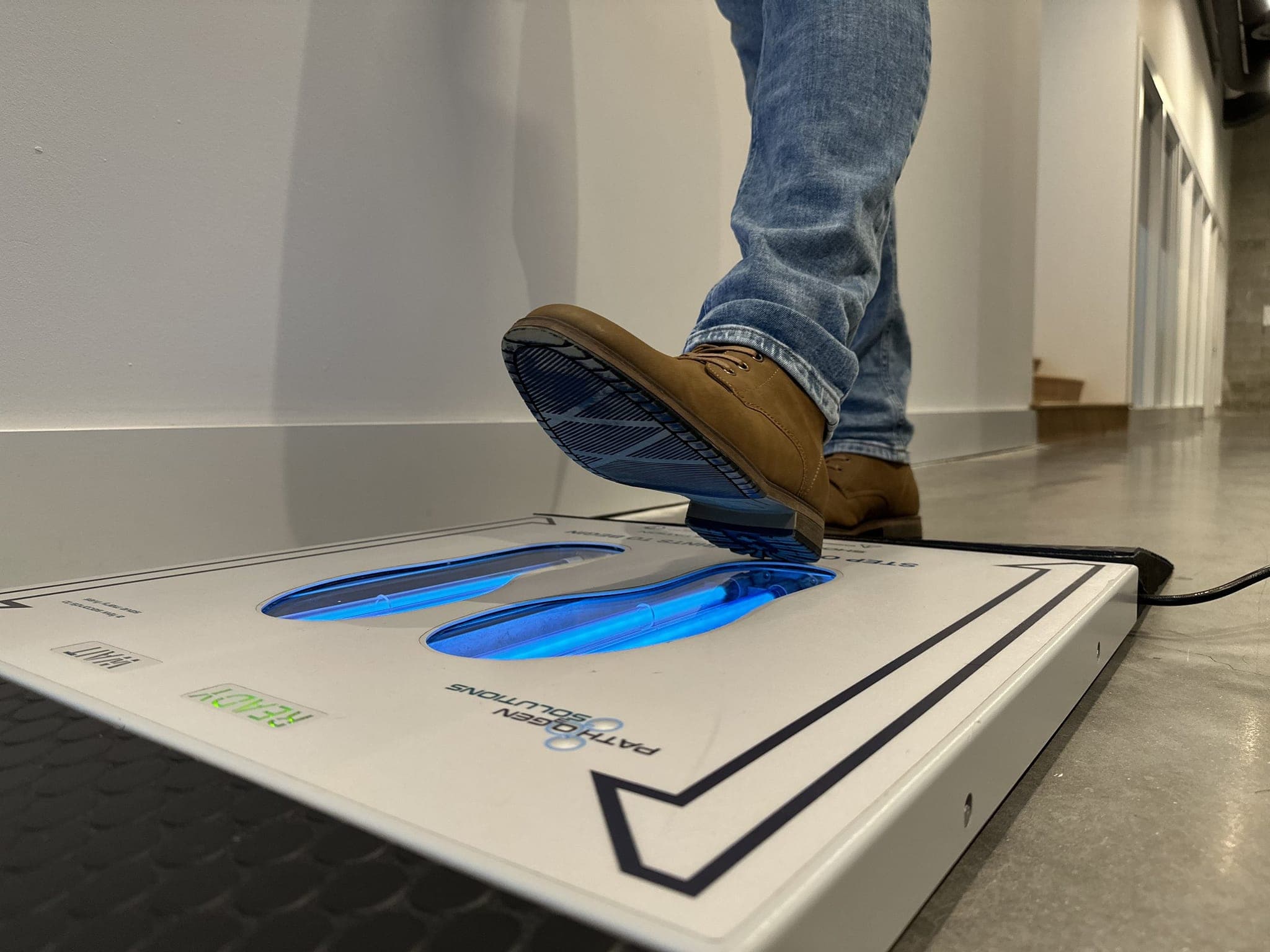 Person in jeans and brown boots stepping onto the UVZone shoe sanitizing station, seen from floor level at the side with the lamps lit