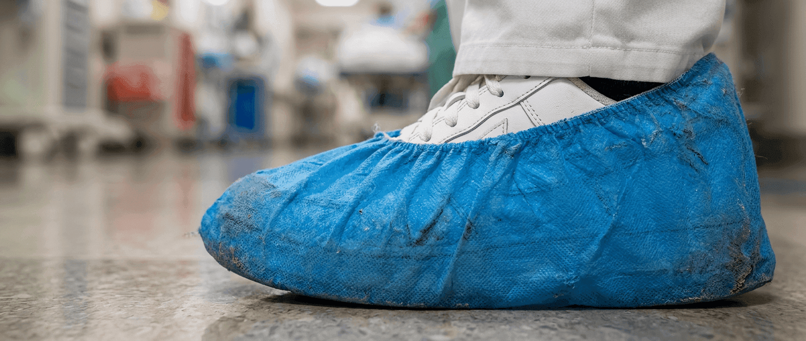 Disposable shoe cover worn over footwear in a clinical corridor, with visible soil on the cover surface.