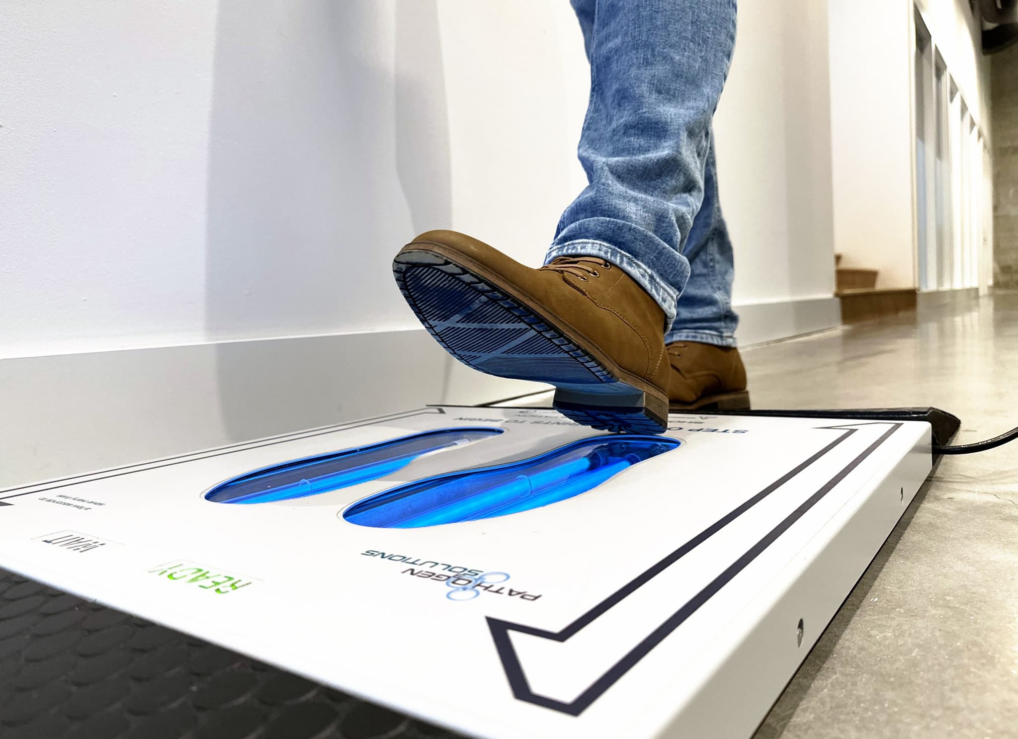 Person in jeans and brown boots stepping onto the UVZone shoe sanitizing station, seen from floor level at the side with the lamps lit