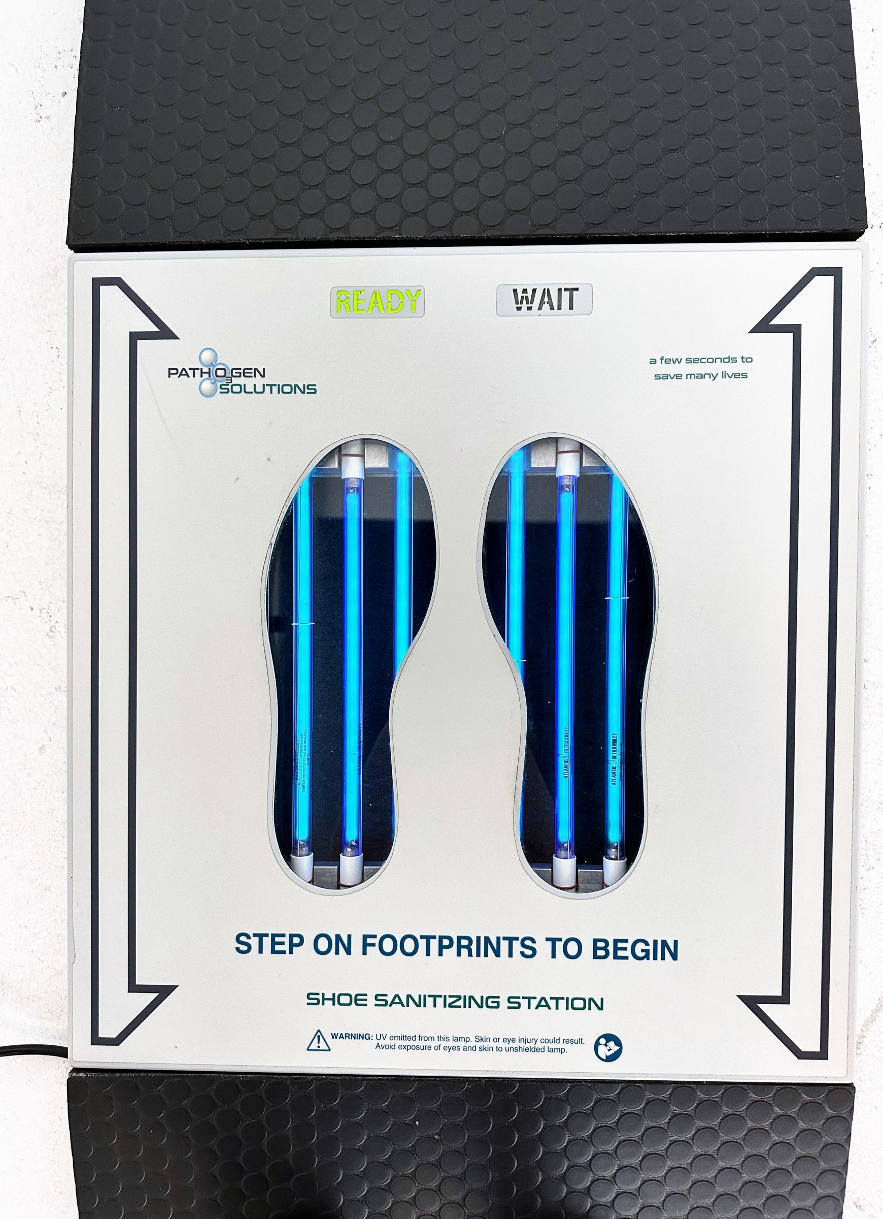 Top of the UVZone shoe sanitizing station showing the READY and WAIT indicators, two footprint windows with lit lamps and the printed instruction to step on footprints to begin